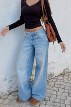 Person wearing a black long-sleeve top and blue jeans standing against a white wall.