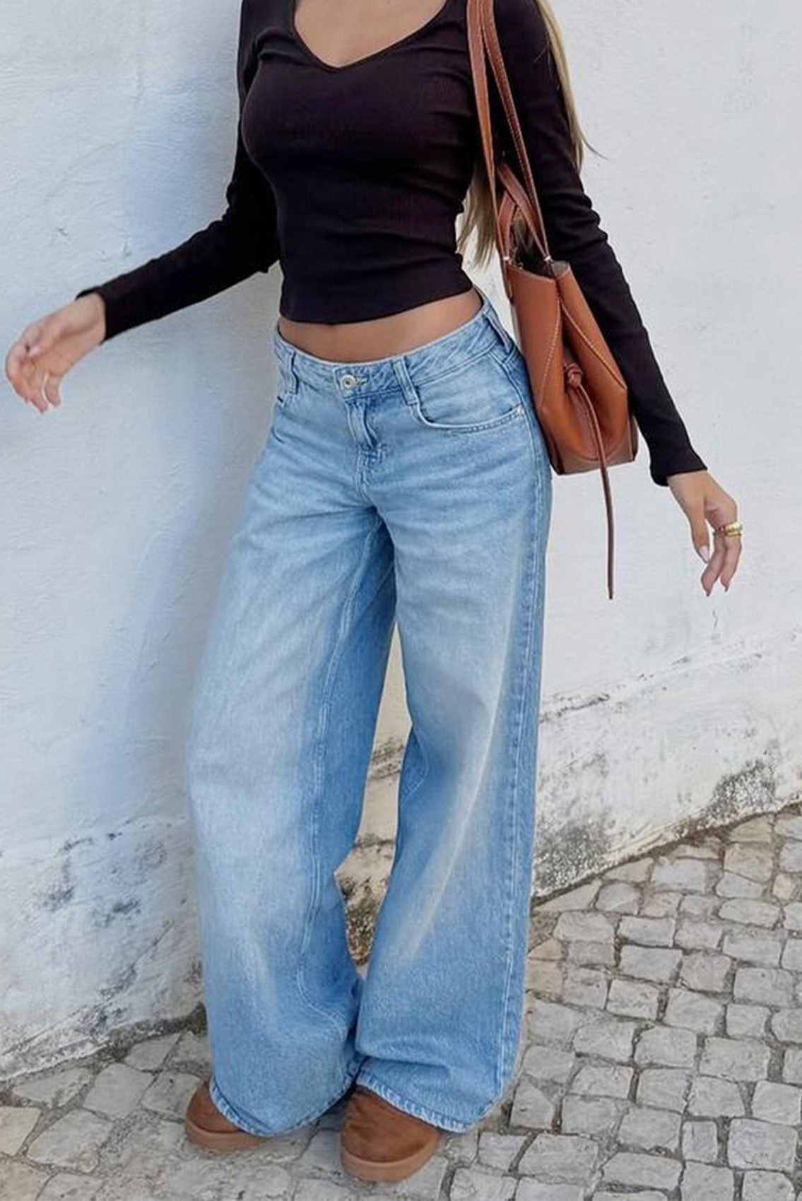 Person wearing a black long-sleeve top and blue jeans standing against a white wall.